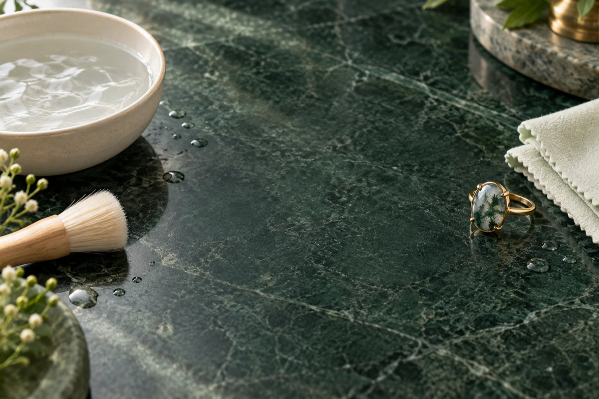 Moss agate ring beside lukewarm water, a soft brush and a lint-free cloth on green stone.