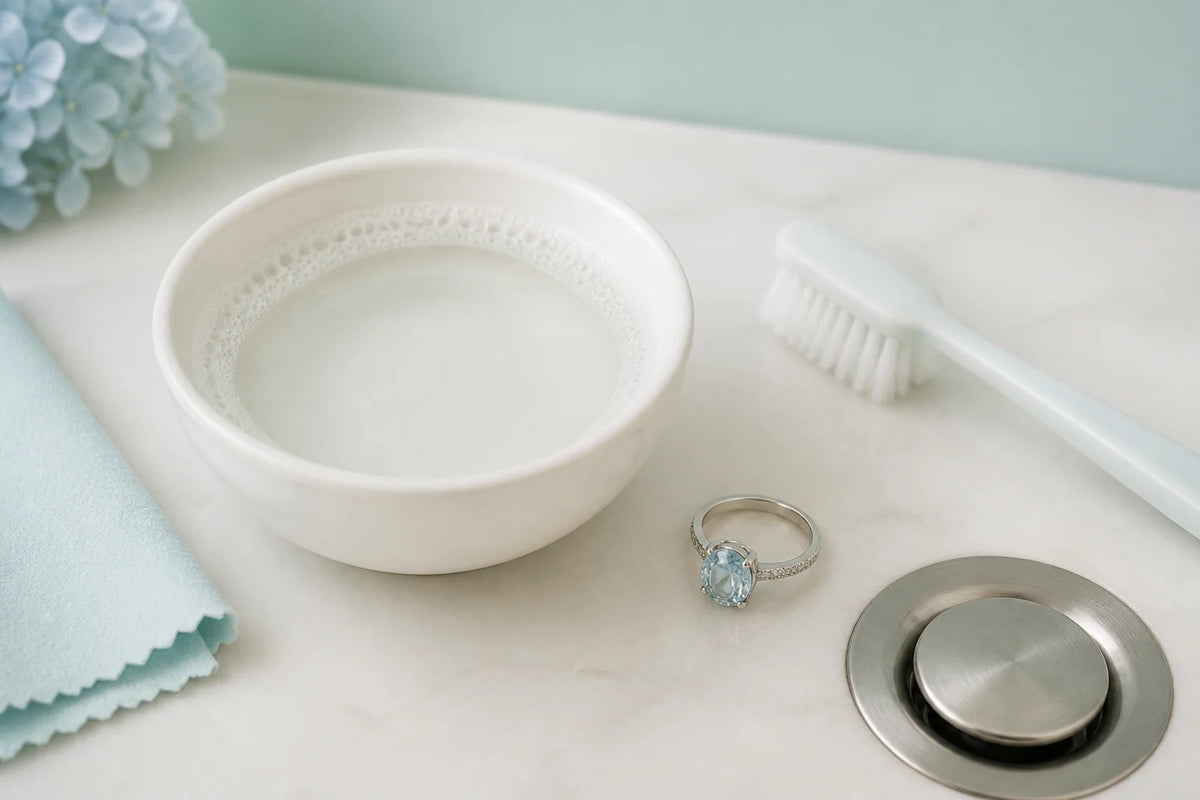 Editorial illustration of an aquamarine ring beside warm soapy water, a soft brush and a lint-free cloth